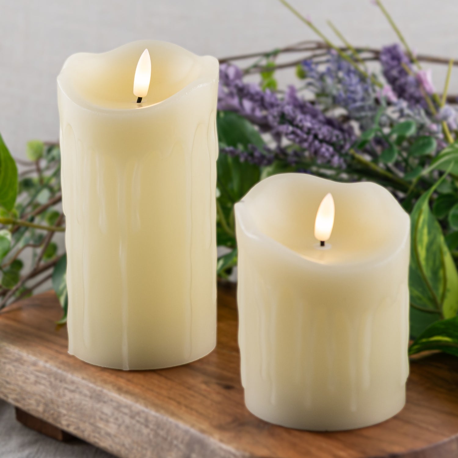 Two flameless candles on a wooden surface with a background of greenery and purple flowers - Picture Perfect Rentals