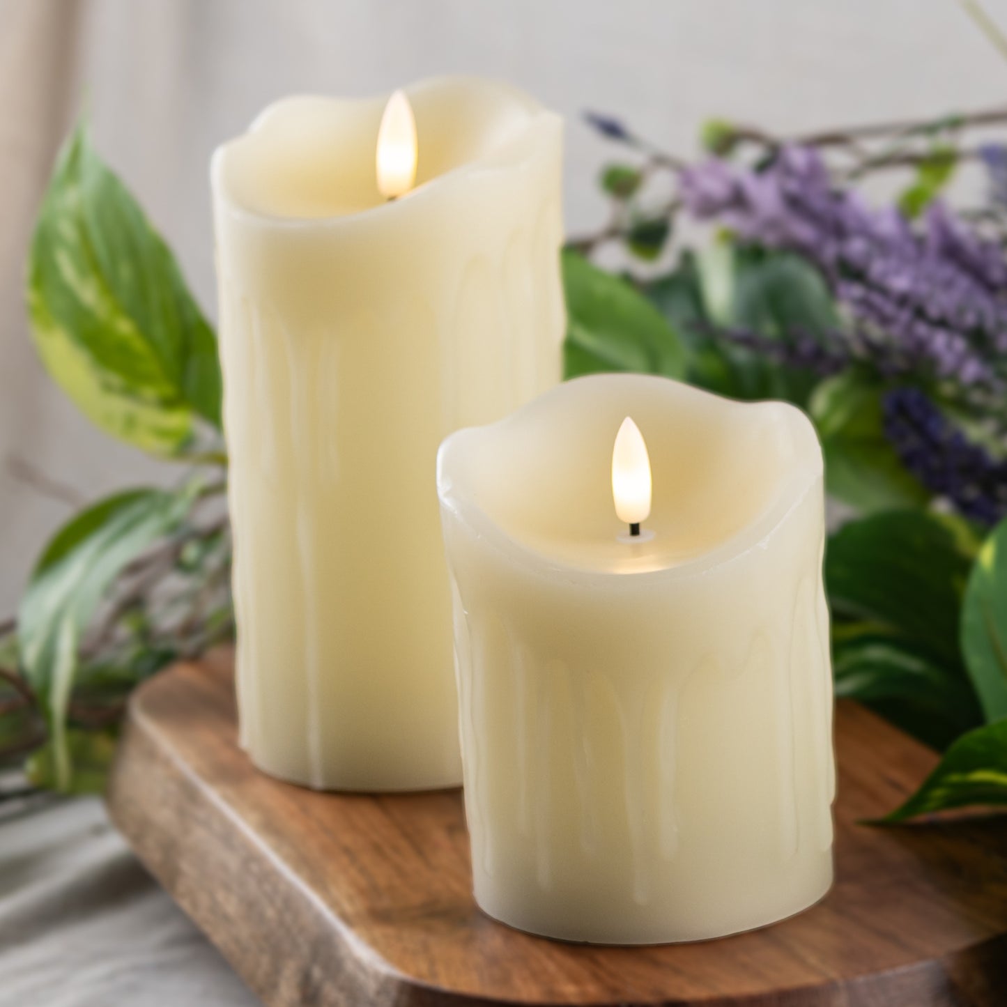 Two flameless candles on a wooden surface with greenery and flowers in the background - Picture Perfect Rentals